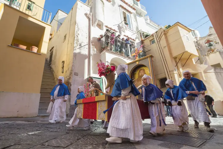 Un gruppo di persone, tra cui la congrega dei turchini, cammina lungo una strada acciottolata a Procida.